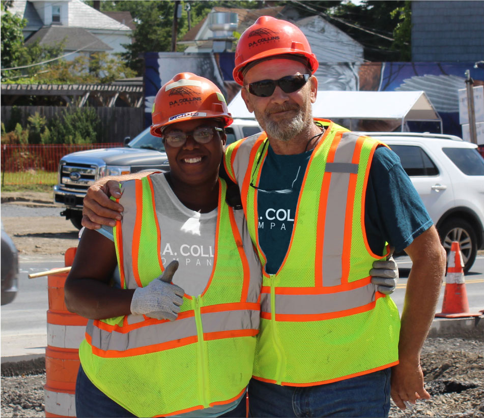 Two D.A. Collins construction workers hugging and smiling. The female on the left is giving a thumbs up